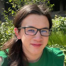 A selfie! I’m smiling at the camera, wearing a green dress, and sitting in front of a large amount of green foliage. It’s a sunny day and shining both on the side of my face and the plants.